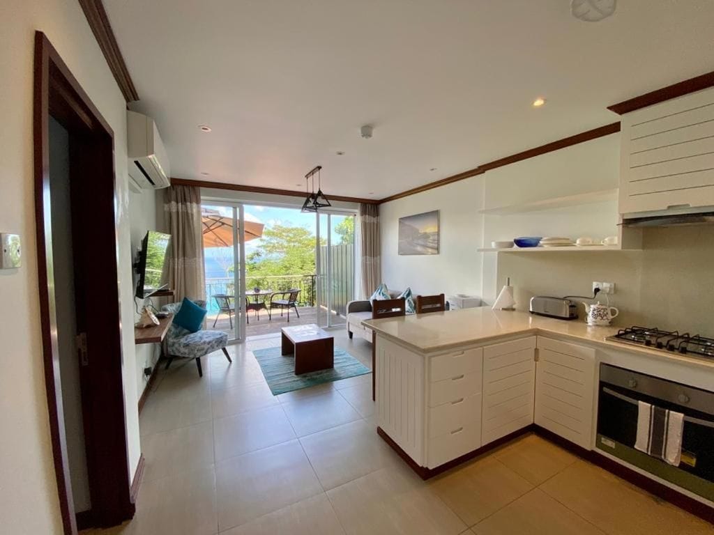 A modern kitchen with a central island, a dining area with a sofa and chairs, and a view through the glass door.