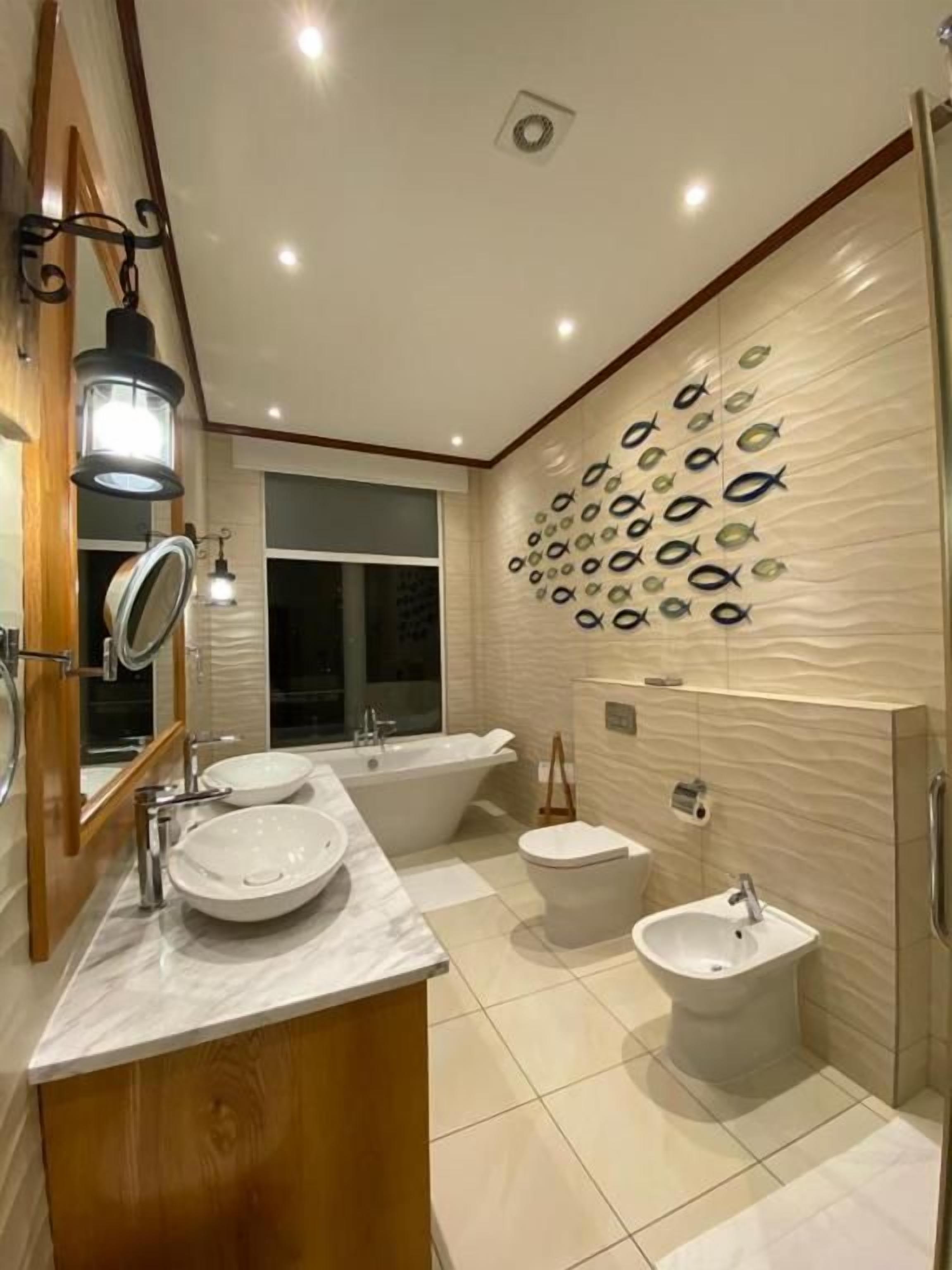 Ein modernes Badezimmer mit einem Doppelwaschtisch, einer freistehenden Badewanne, einem Bidet und einer Wanddekoration mit Fischen.
