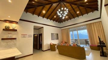 A spacious living area with a high ceiling and a central chandelier, a seating area with a sofa and chairs, and a view through a large window.