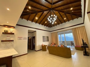 A spacious living area with a high ceiling and a central chandelier, a seating area with a sofa and chairs, and a view through a large window.