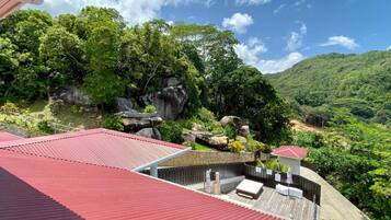 A rooftop terrace with a red roof, surrounded by lush greenery and a mountainous landscape.