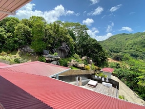 A rooftop terrace with a red roof, surrounded by lush greenery and a mountainous landscape.