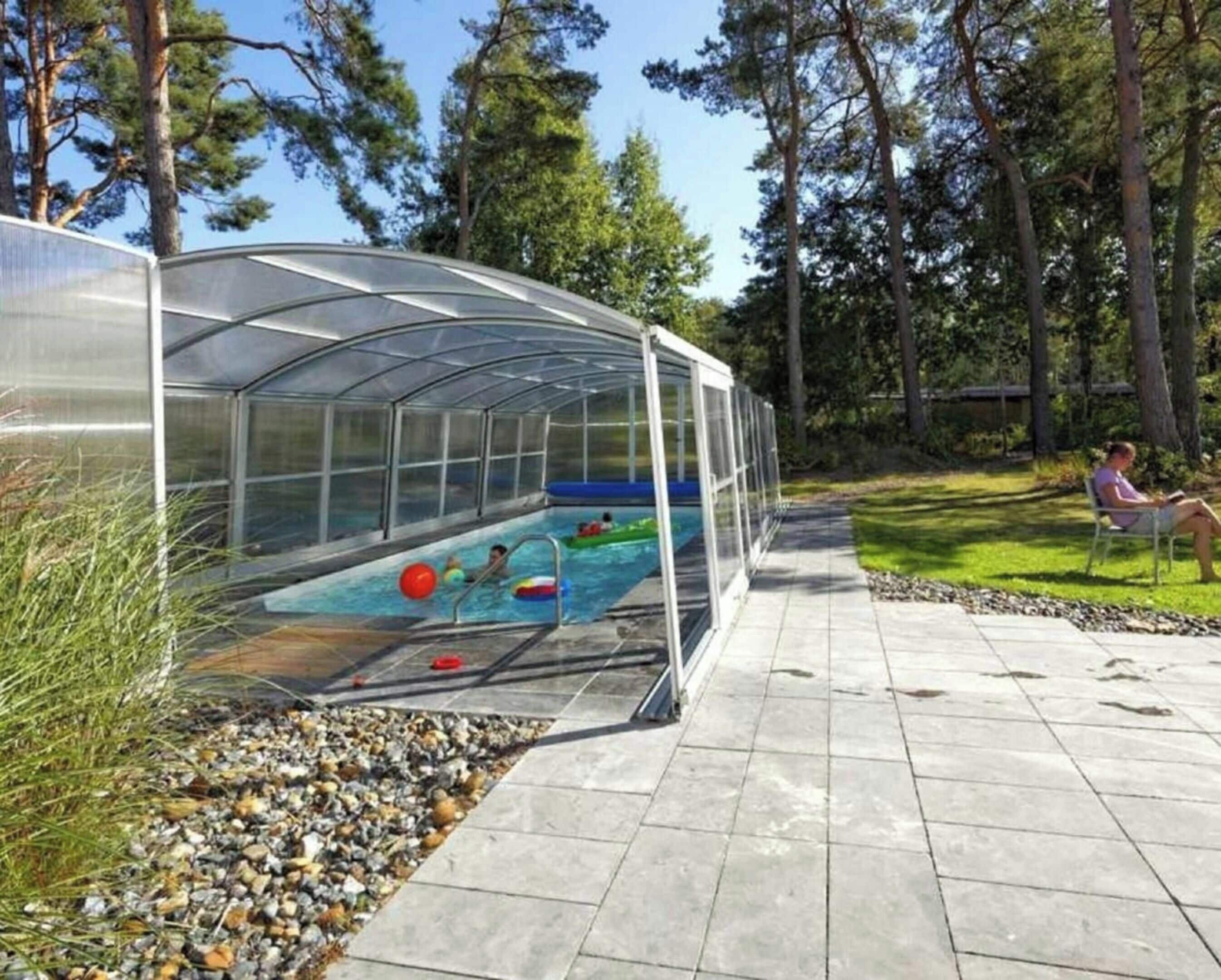 Une piscine couverte, protégée par une structure en verre, entourée d’un bord en galets et d’un chemin pavé.