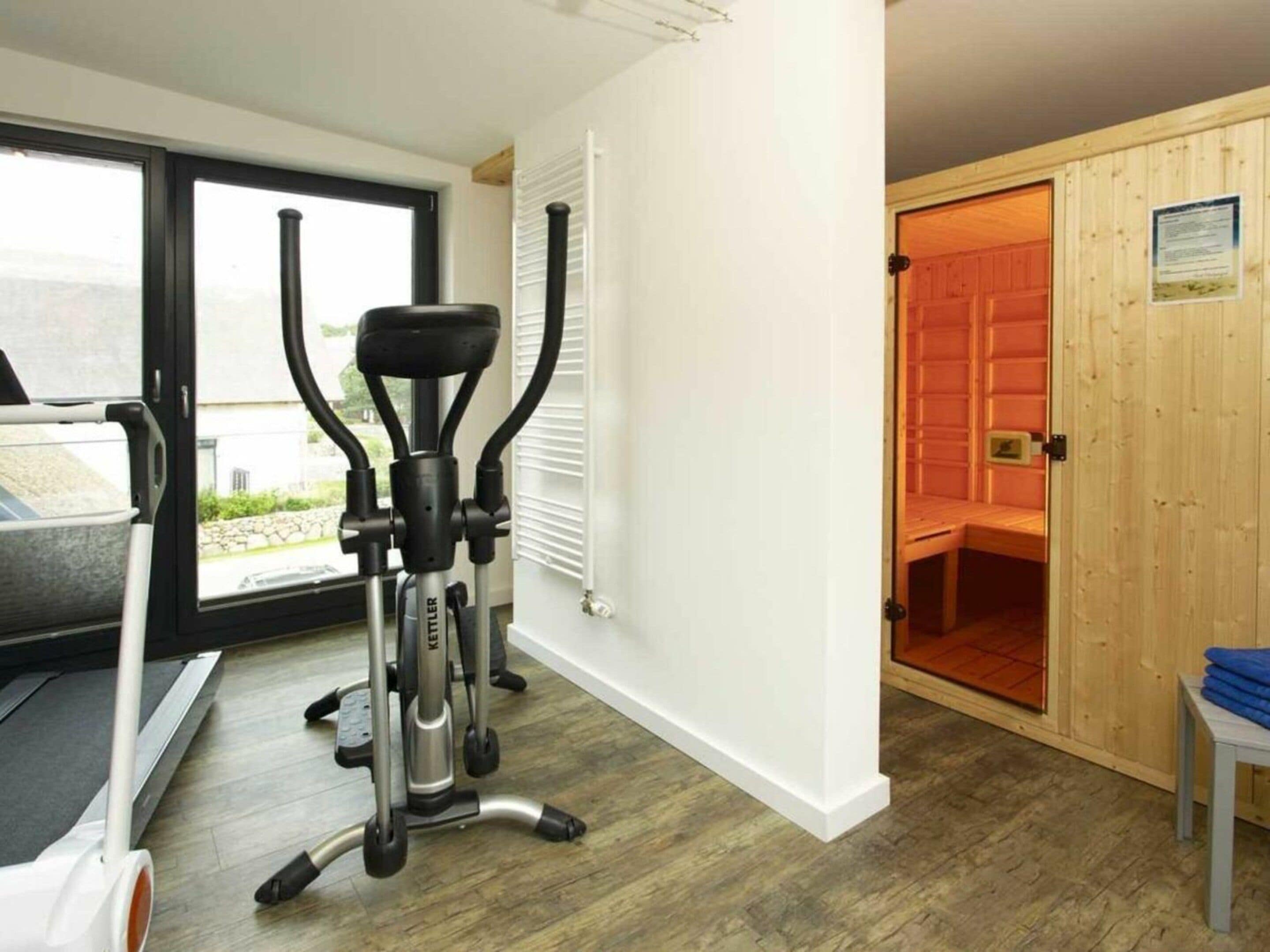 A room with a treadmill, exercise equipment, a wooden door leading to a sauna, and a view through a glass door.