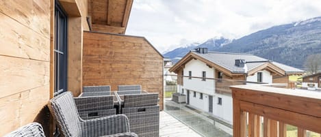 Una terraza de madera con mobiliario de mimbre, barandilla de vidrio y vista a un paisaje montañoso.