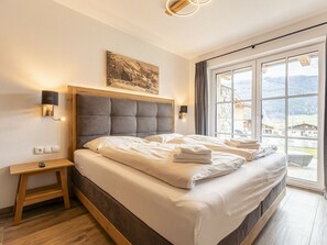 A hotel room with a large bed, a bedside table, a wall-mounted light, a painting, and a window with a mountain view. - Superior Holiday Home # 5A With Sauna (Uttendorf)