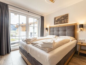 Una cama doble con cabecera de madera, una mesita de noche con lámpara, y vistas a un paisaje montañoso a través de amplios ventanales.