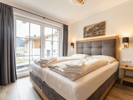 Una cama doble con cabecera de madera, una mesita de noche con lámpara, y vistas a un paisaje montañoso a través de amplios ventanales.
