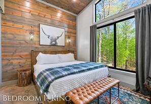 Ein Schlafzimmer mit einer Holzwand, einem Bett mit weißer Bettwäsche, einer Bank, einem Fenster mit Blick auf Bäume und einem gerahmten Bild an der Wand.
