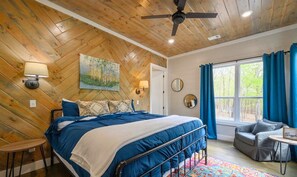 Ein Schlafzimmer mit Zargenholzboden, einem Bett mit blauer Bettwäsche, einem kleinen Tisch, einem Stuhl und einem Fenster mit Vorhängen.