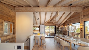 A modern kitchen with wooden beams, a dining area with a long table, and a view of the outdoors. - Chalet Montâme - Modern luxury in the Swiss Alps (Riddes)
