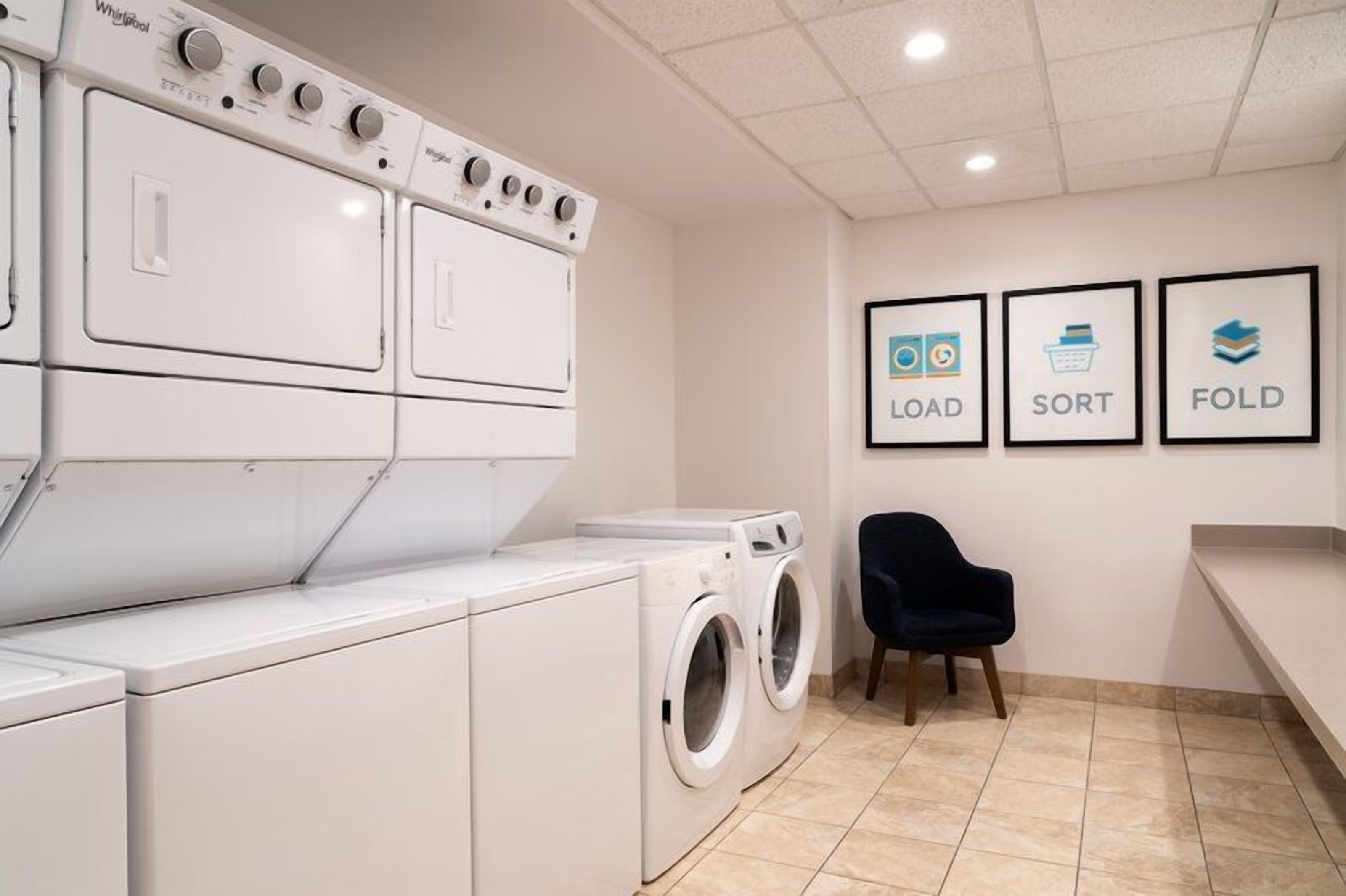 A laundry room with stacked washers and dryers, a chair, and wall-mounted signs indicating 'LOAD,' 'SORT,' and 'FOLD' functions.