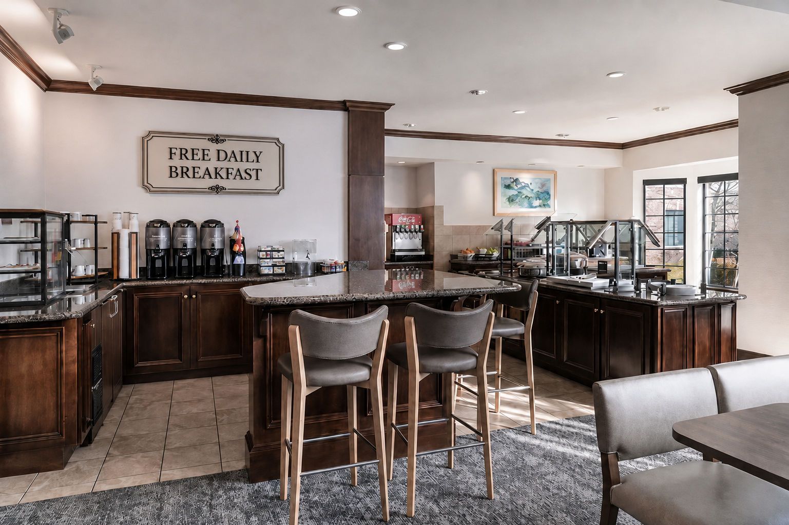A modern kitchen with a bar area, a sign indicating free daily breakfast, and a large window providing natural light.