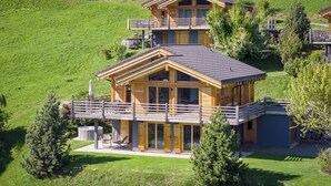 A two-story wooden cabin with a balcony, surrounded by a grassy landscape. - Chalet Aramis - Four Bedroom Chalet, Sleeps 8 (Riddes)