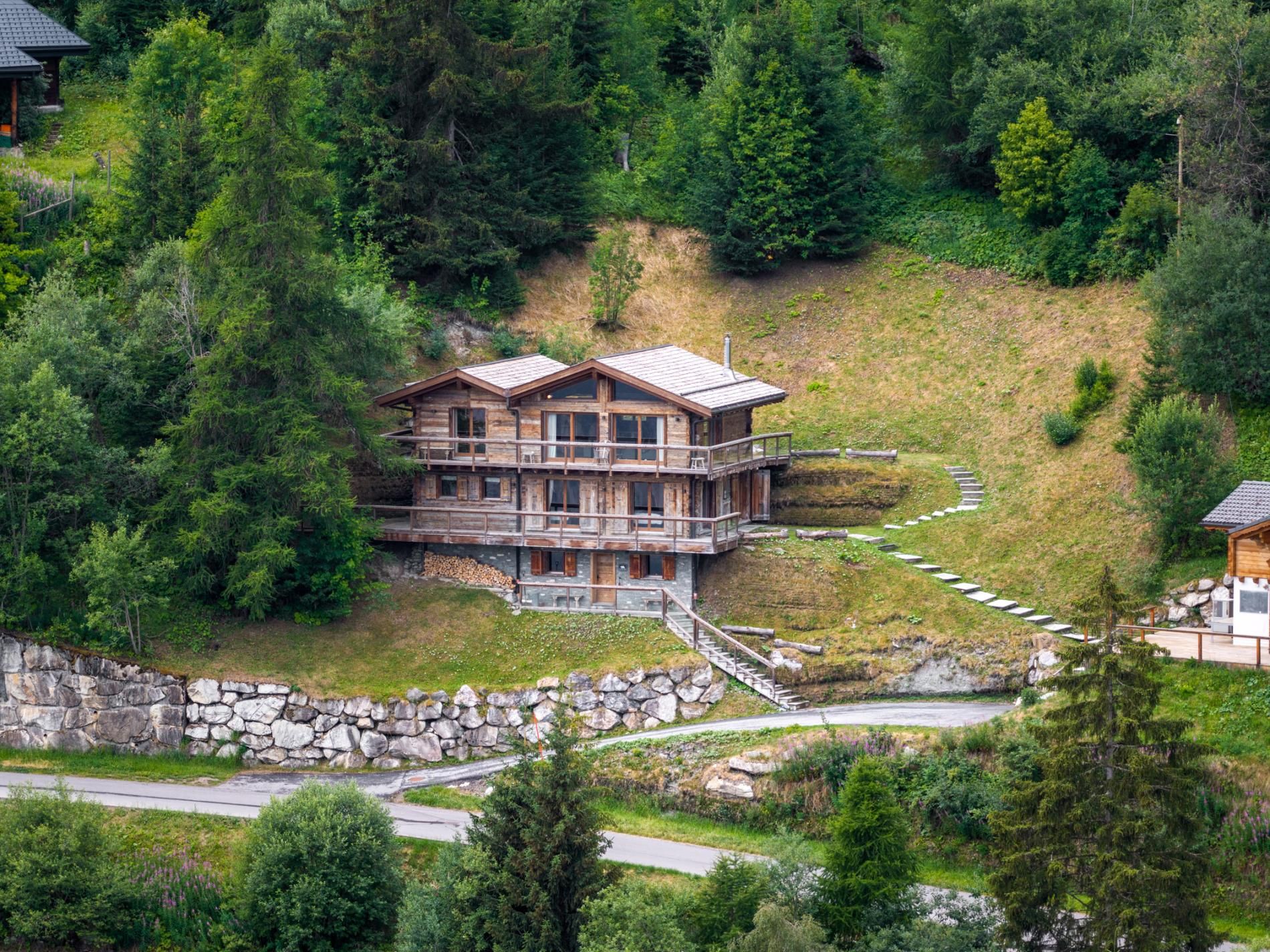 A multi-story wooden cabin with a balcony, surrounded by dense forest and a stone retaining wall.