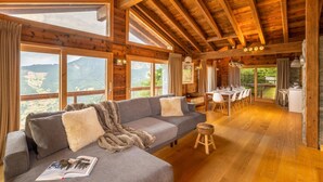 Interno di una baita in legno con divano, tavolo da pranzo e sedie, ampie finestre con vista sulle montagne.