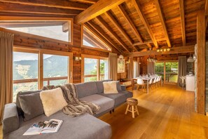 A wooden cabin interior with a sofa, dining table, and chairs, large windows with a mountain view. - Chalet Big Bear - Five Bedroom Chalet, Sleeps 10 (Riddes)
