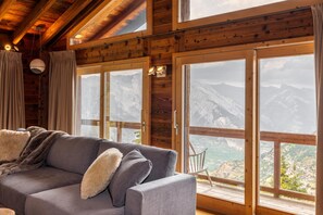 Ein gemütliches Zimmer mit grauem Sofa, Holzbalken und großen Fenstern mit Blick auf die Berge.