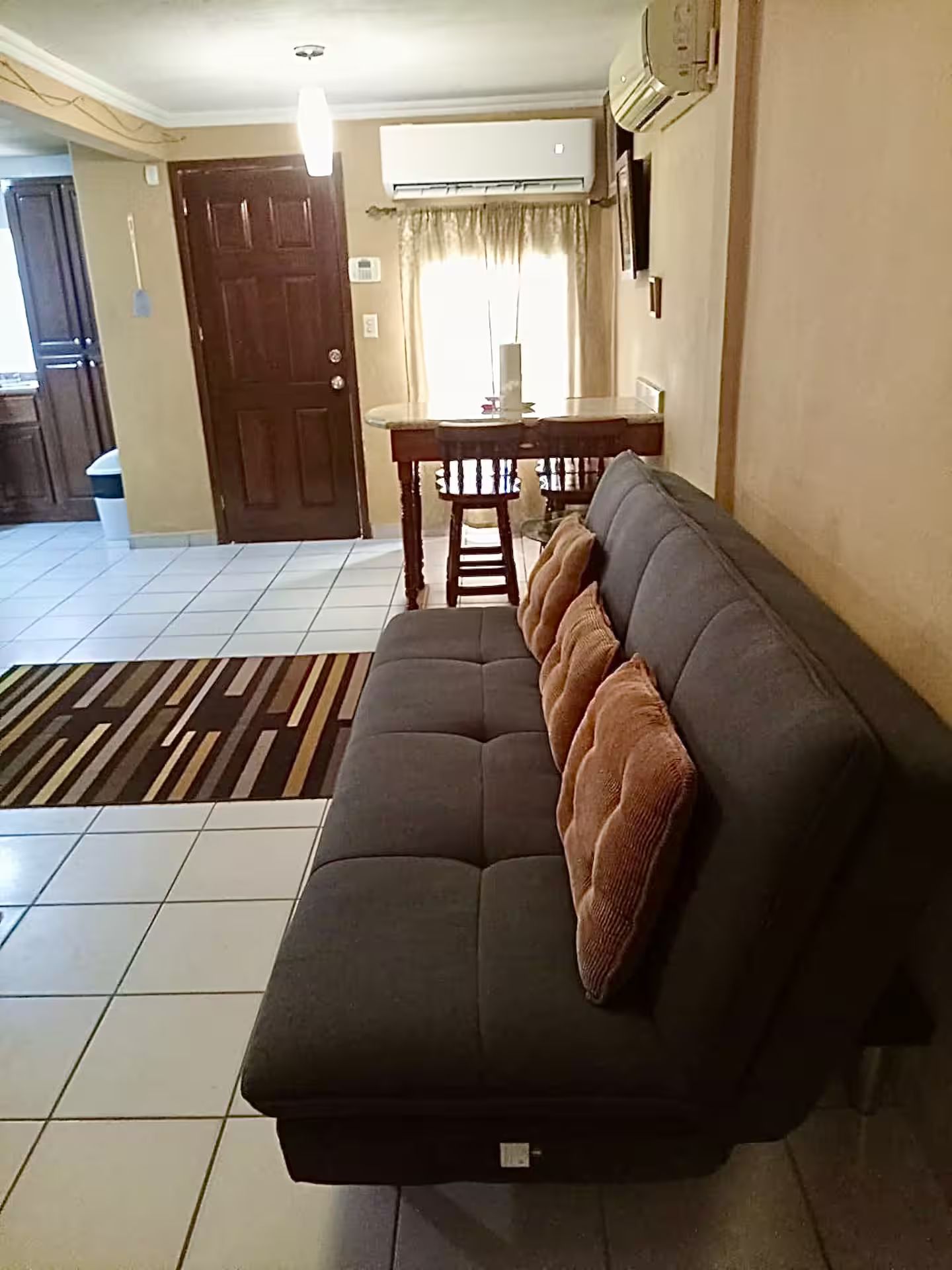 Un salon avec un canapé foncé, un tapis à motifs, une table à manger avec des chaises, et un coin cuisine à l’arrière-plan.