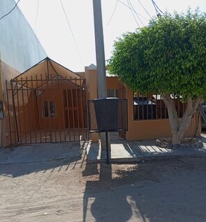 A gated entrance with a metal gate, a mailbox, and a tree in front of a building with a beige facade. - Brisas del Golfo Little House (Puerto Peñasco)
