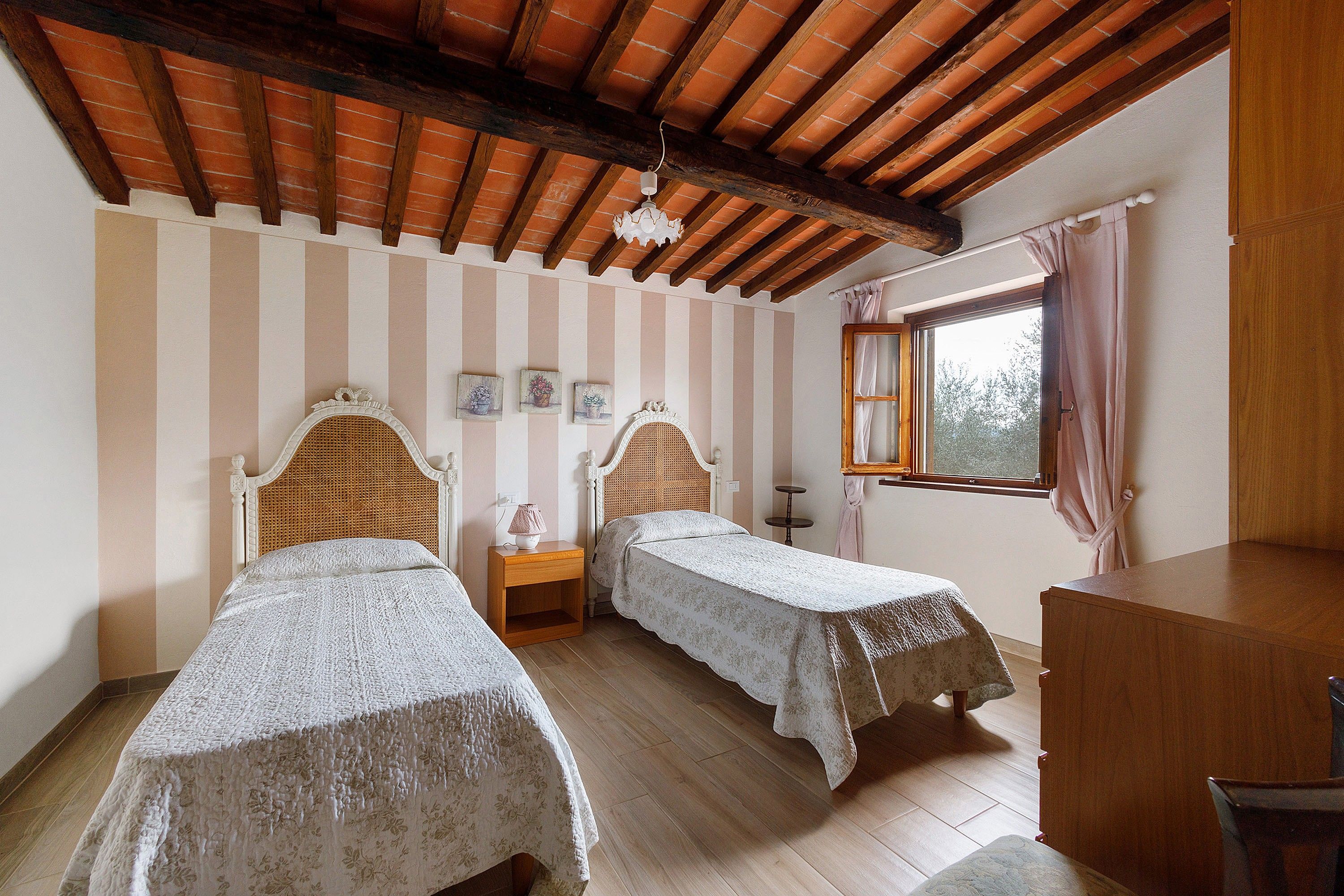 A room with two beds, a wooden ceiling, a window with curtains, and a small table.