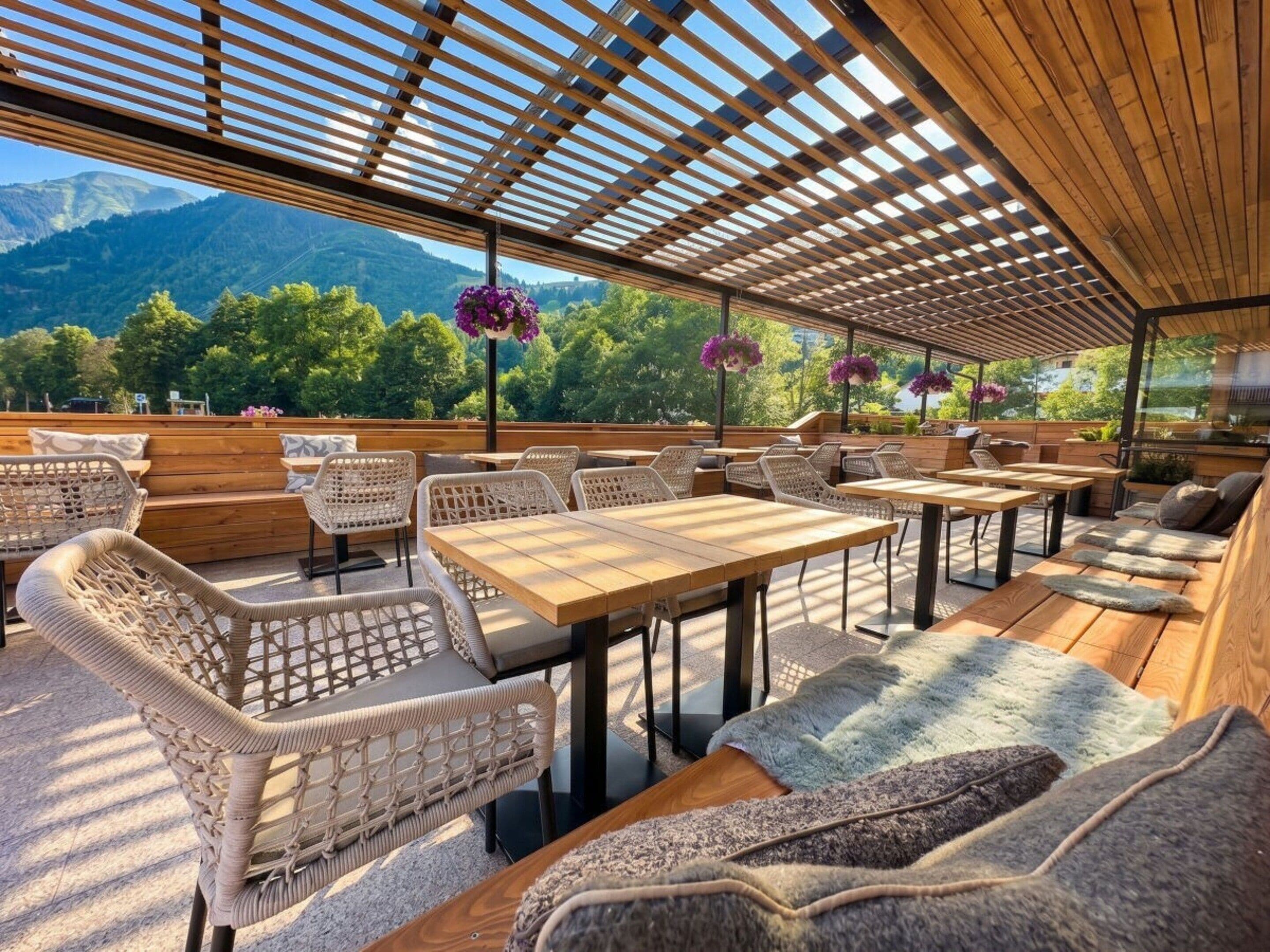 Un área de estar al aire libre con mesas y sillas de madera, una pérgola en lo alto y vistas a montañas al fondo.