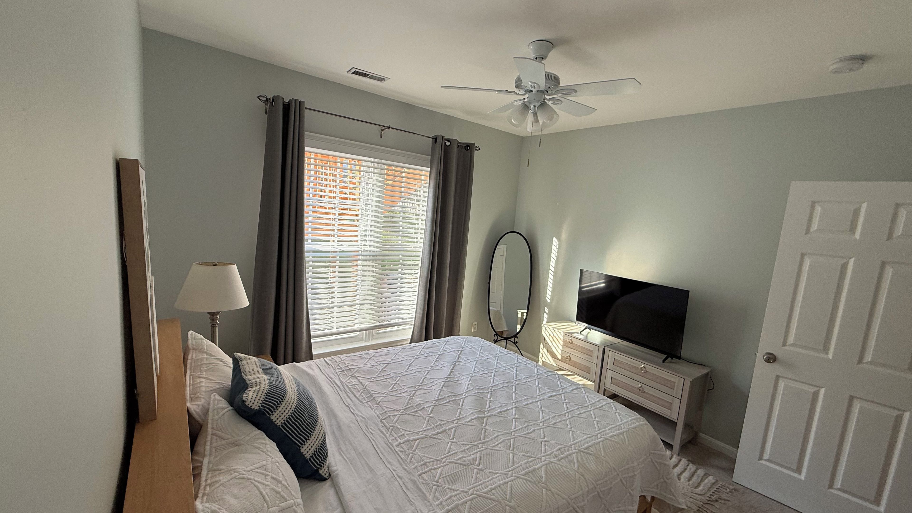 A bedroom with a bed, a TV, a dresser, a ceiling fan, and a window with curtains.