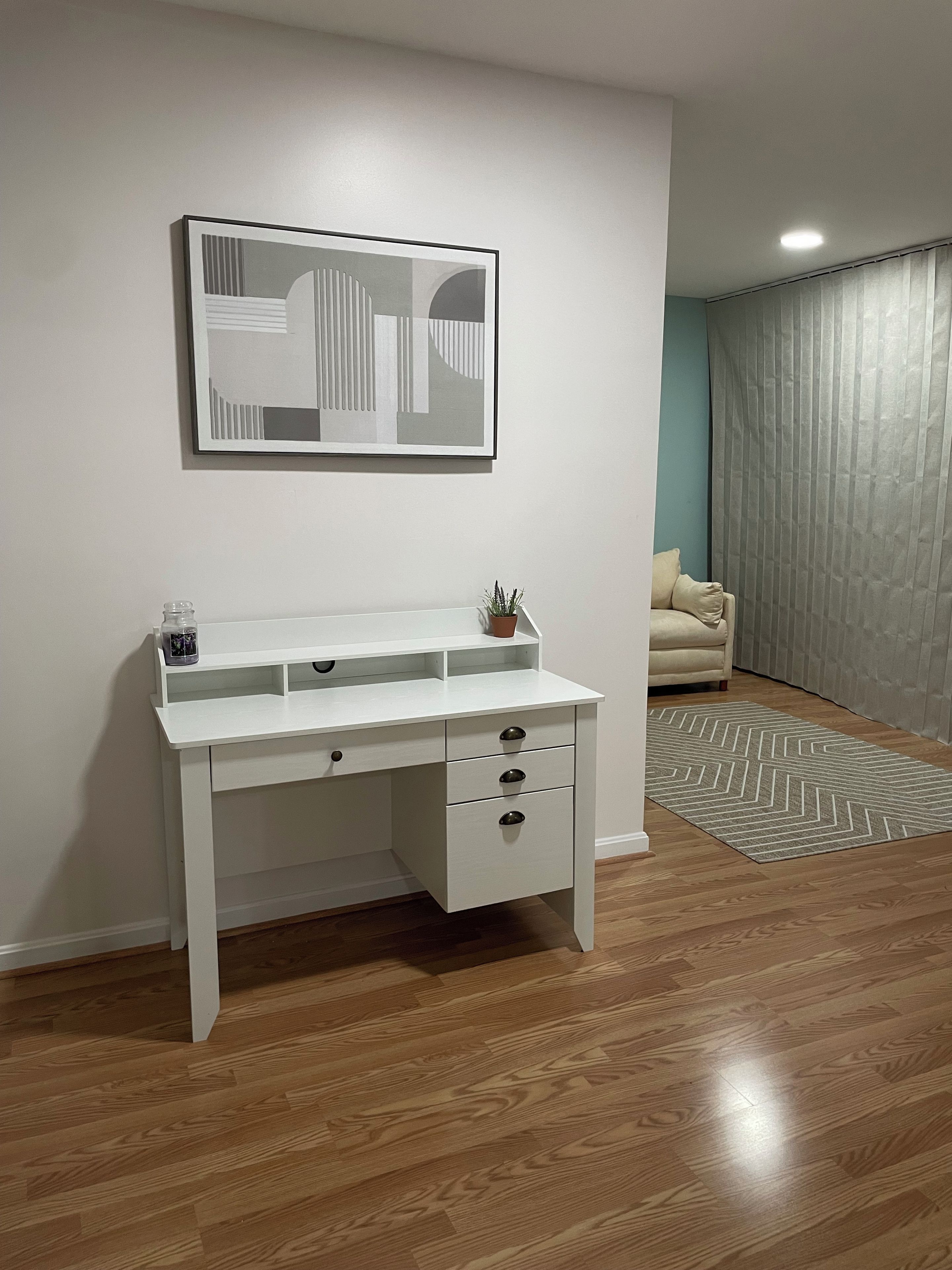 A white desk with drawers and a shelf, a framed abstract artwork on the wall, a potted plant, and a patterned rug on the floor.