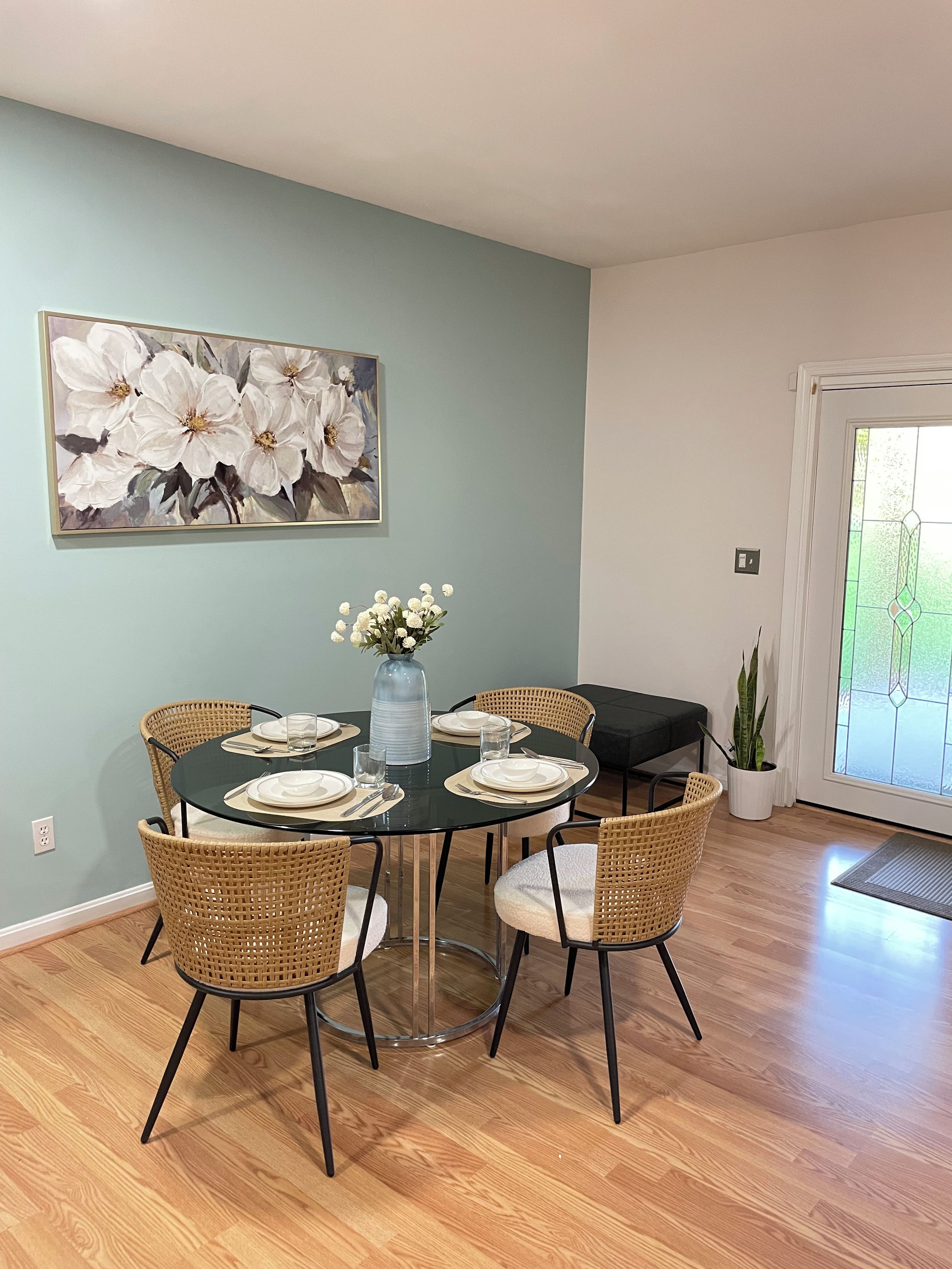 A dining area with a round table set for four, surrounded by wicker chairs, a vase with flowers, and a large floral painting on the wall.