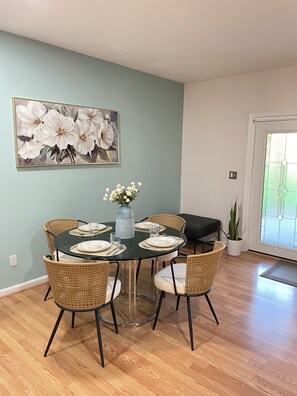 Een eetruimte met een ronde tafel voor vier personen, omringd door rieten stoelen, een vaas met bloemen en een groot bloemschilderij aan de muur.