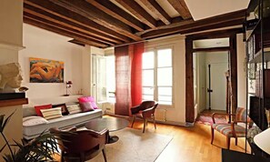 A room with a wooden ceiling, a sofa with cushions, a dining table, chairs, a window with curtains, and a bookshelf. - BEAUTIFUL 2-ROOM APARTMENT IN THE HEART OF THE PRESTIGIOUS 6TH ARRONDISSEMENT (Paris)