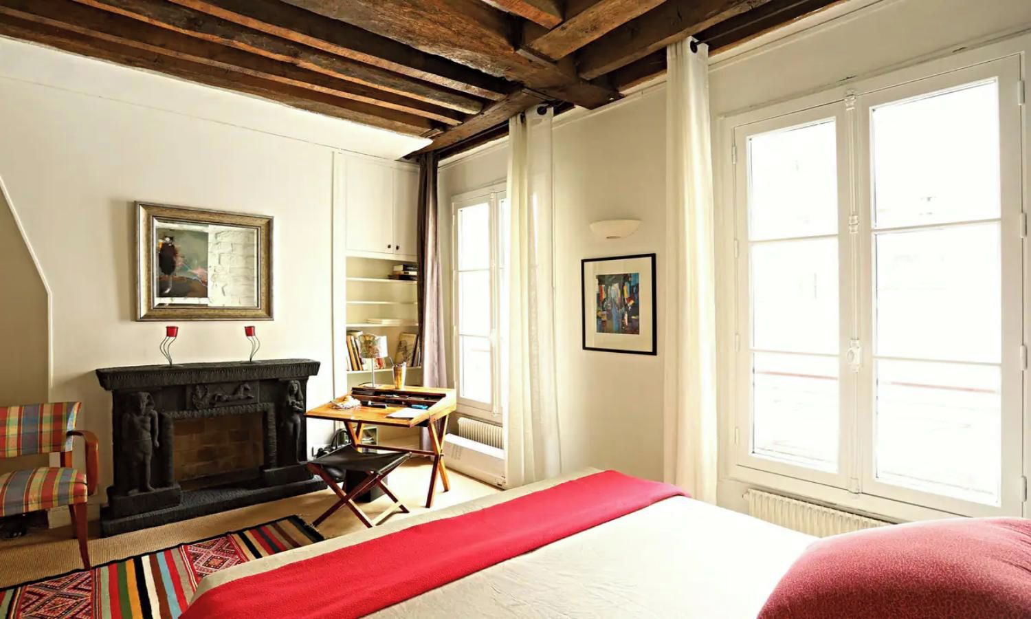 Una habitación con chimenea, una cama con una manta roja, un escritorio y una estantería.