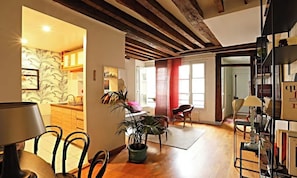 A spacious living area with a wooden ceiling, a dining table with chairs, a bookshelf, and a large window with curtains. - BEAUTIFUL 2-ROOM APARTMENT IN THE HEART OF THE PRESTIGIOUS 6TH ARRONDISSEMENT (Paris)