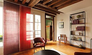 A room with a wooden floor, a red chair, a black table, a bookshelf, and a window with curtains. - BEAUTIFUL 2-ROOM APARTMENT IN THE HEART OF THE PRESTIGIOUS 6TH ARRONDISSEMENT (Paris)