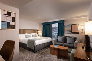 A hotel room with a bed, a sofa, a desk, and a chair. There is a bulletin board with various notices on the wall. - Newcastle SuperKing Studio Suite | Free Breakfast! (Newcastle Upon Tyne)