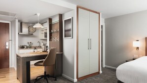 Ein Hotelzimmer mit einer Küchenzeile, einem Bett, einem Schreibtisch mit Stuhl, einem Kleiderschrank und einem Nachttisch mit Lampe.