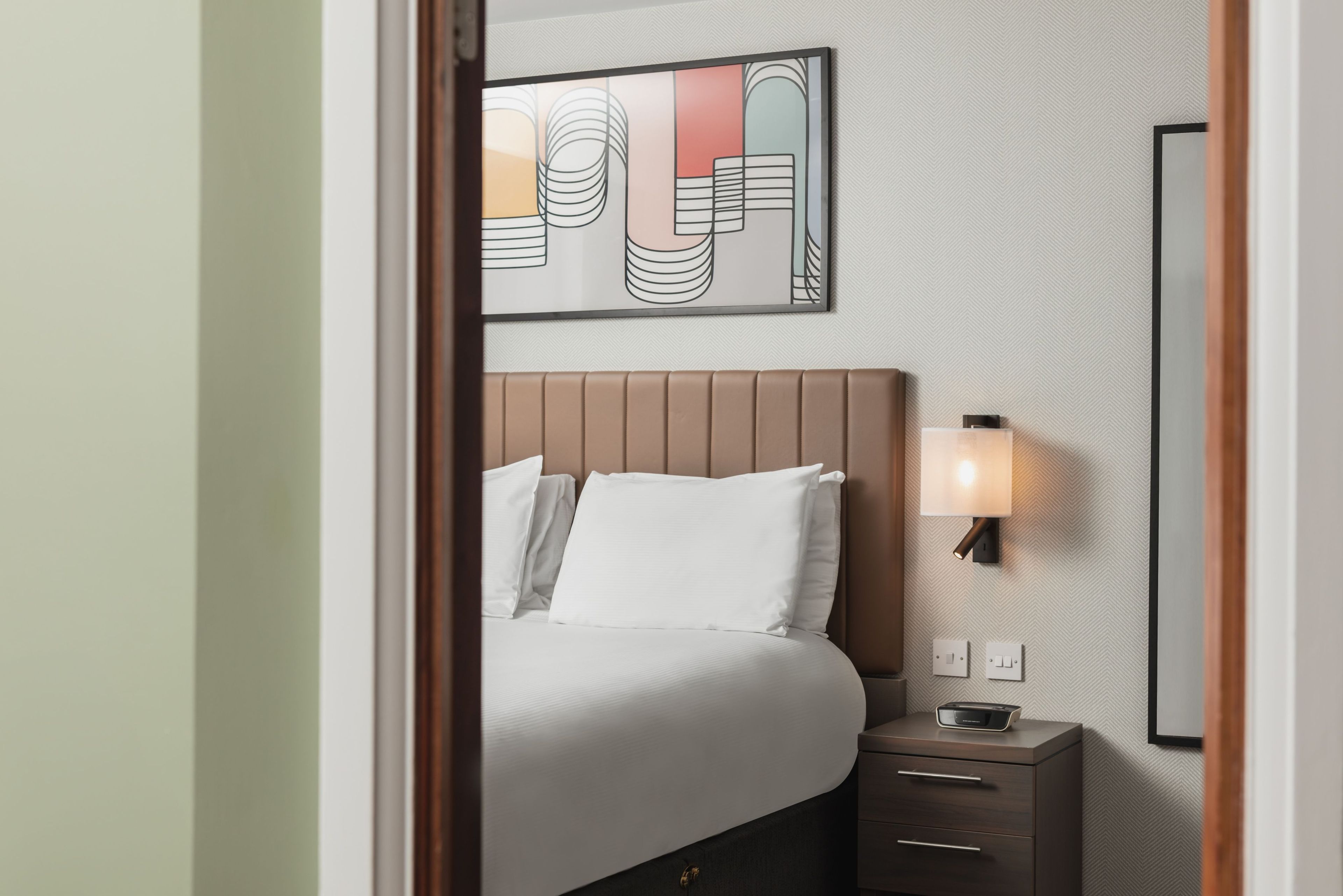 Una habitación de hotel con cama, mesita de noche, lámpara y una obra de arte colgada en la pared.