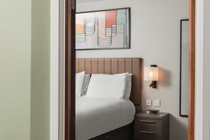 A hotel room with a bed, bedside table, lamp, and a wall-mounted artwork. - Suite w/ Living Area + Kitchen | Free Breakfast! (Newcastle Upon Tyne)