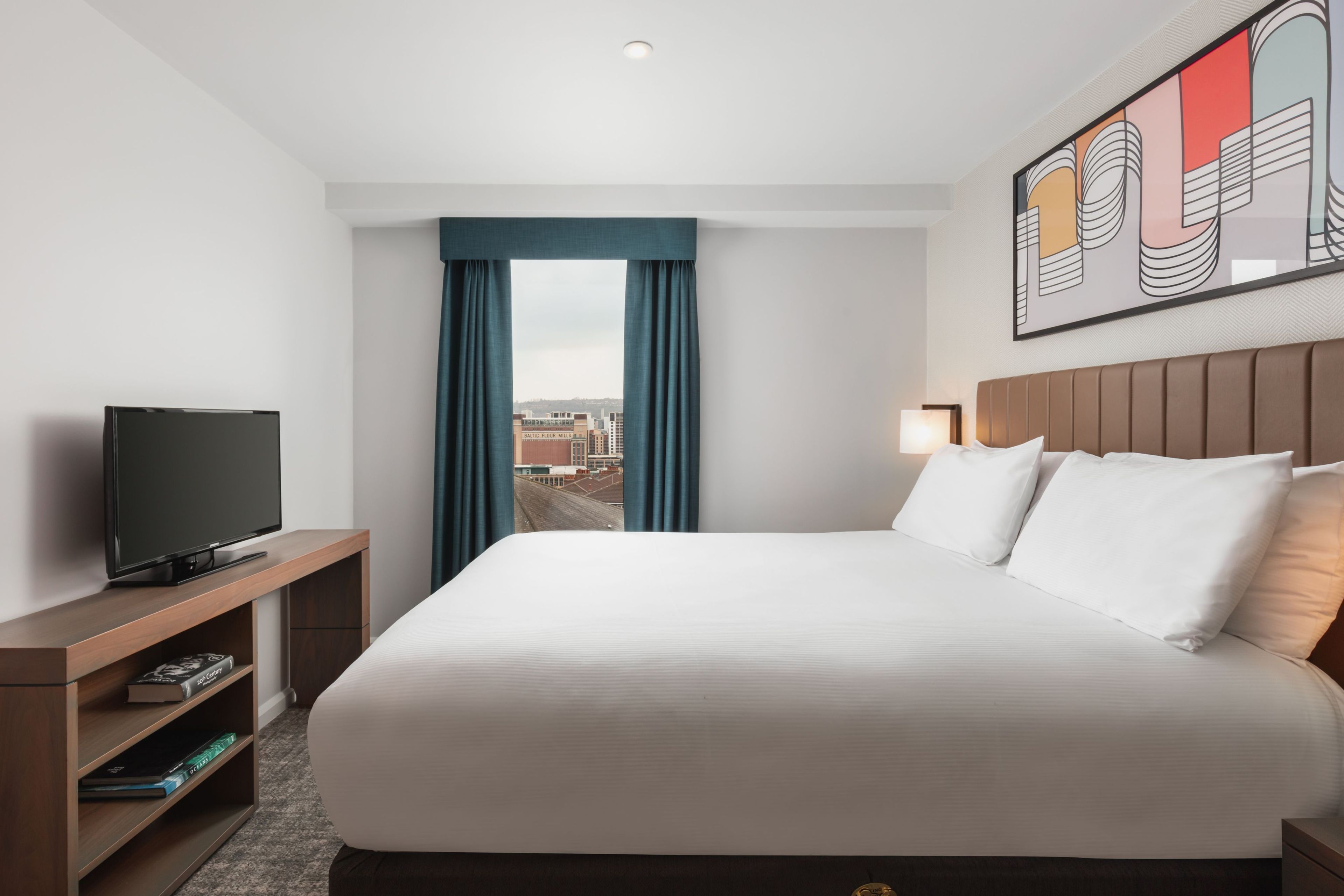 Ein Hotelzimmer mit einem großen Bett, einem Flachbildfernseher auf einem Holzboden, Blick auf die Stadt durch ein Fenster mit blauen Vorhängen und einem modernen Kunstwerk an der Wand.