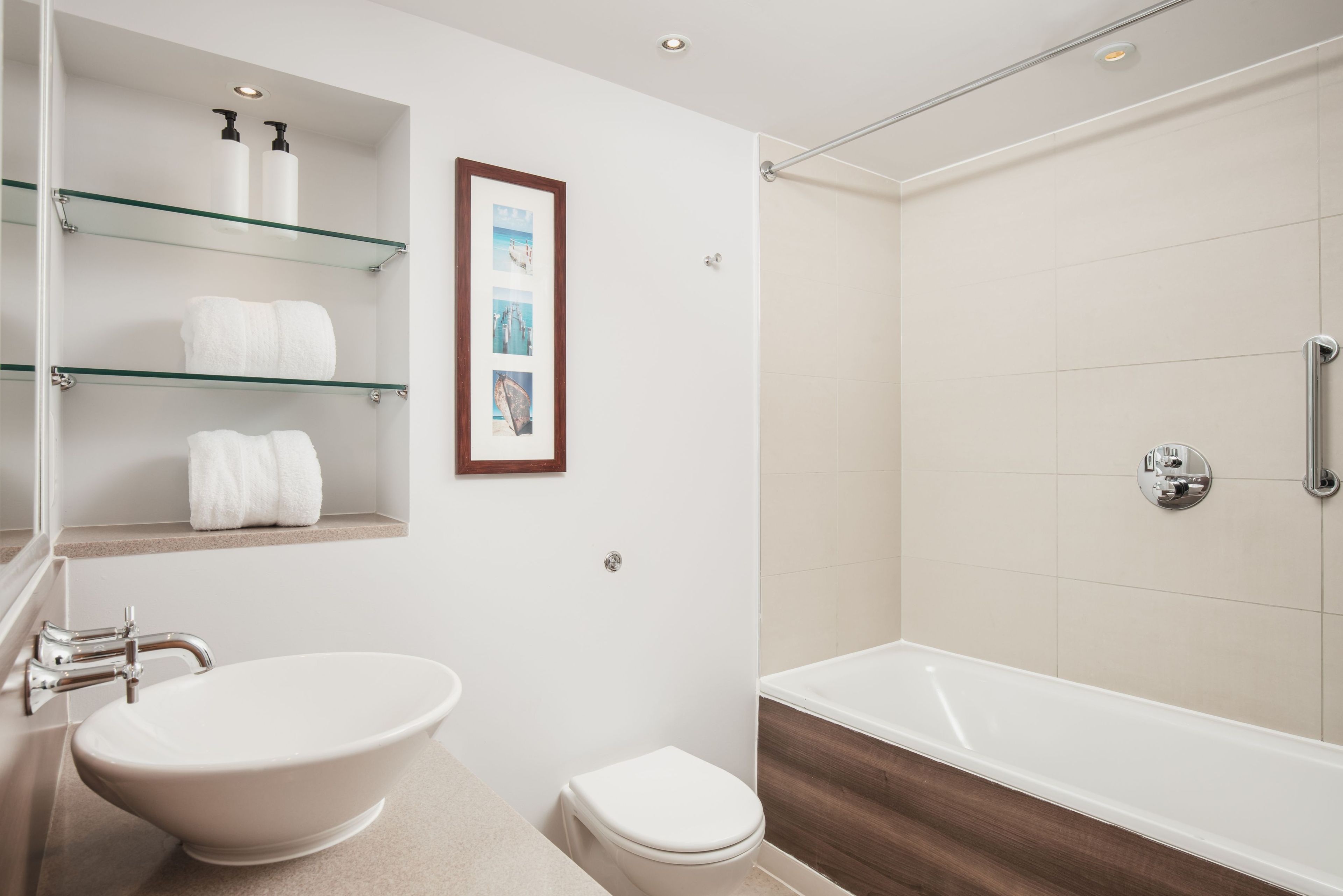 A bathroom with a freestanding bathtub, a sink with a vessel basin, and a toilet. There are glass shelves with toiletries and a framed picture on the wall.