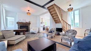 A spacious living room with a fireplace, a staircase, and a kitchen in the background. - Charming 2-bedroom house in the Baie de Somme (Noyelles-sur-Mer)