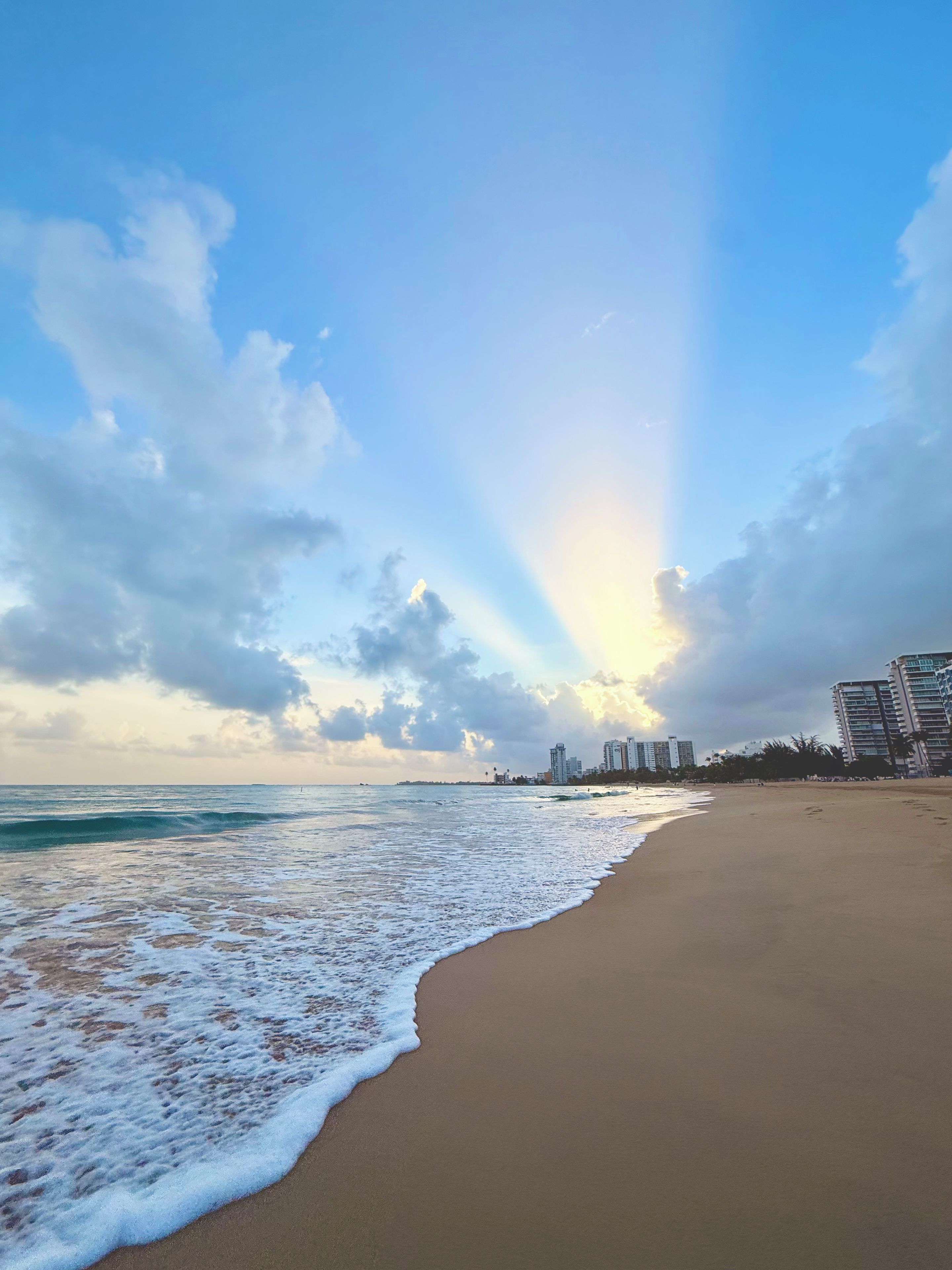 A beach with a clear sky, waves, and buildings in the distance.
