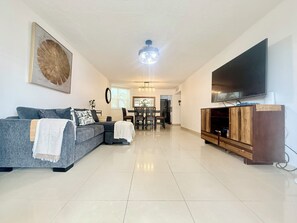 A modern living room with a grey sectional sofa, a flat-screen TV mounted on the wall, and a wooden entertainment unit. - Brisa Caribe Marbella Elegant 3BR Retreat Steps from Beach in Isla Verde, PR!! (Carolina)