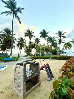 A beach playground with swings and a slide, surrounded by palm trees and a sandy area. - Brisa Caribe Marbella Elegant 3BR Retreat Steps from Beach in Isla Verde, PR!! (Carolina)