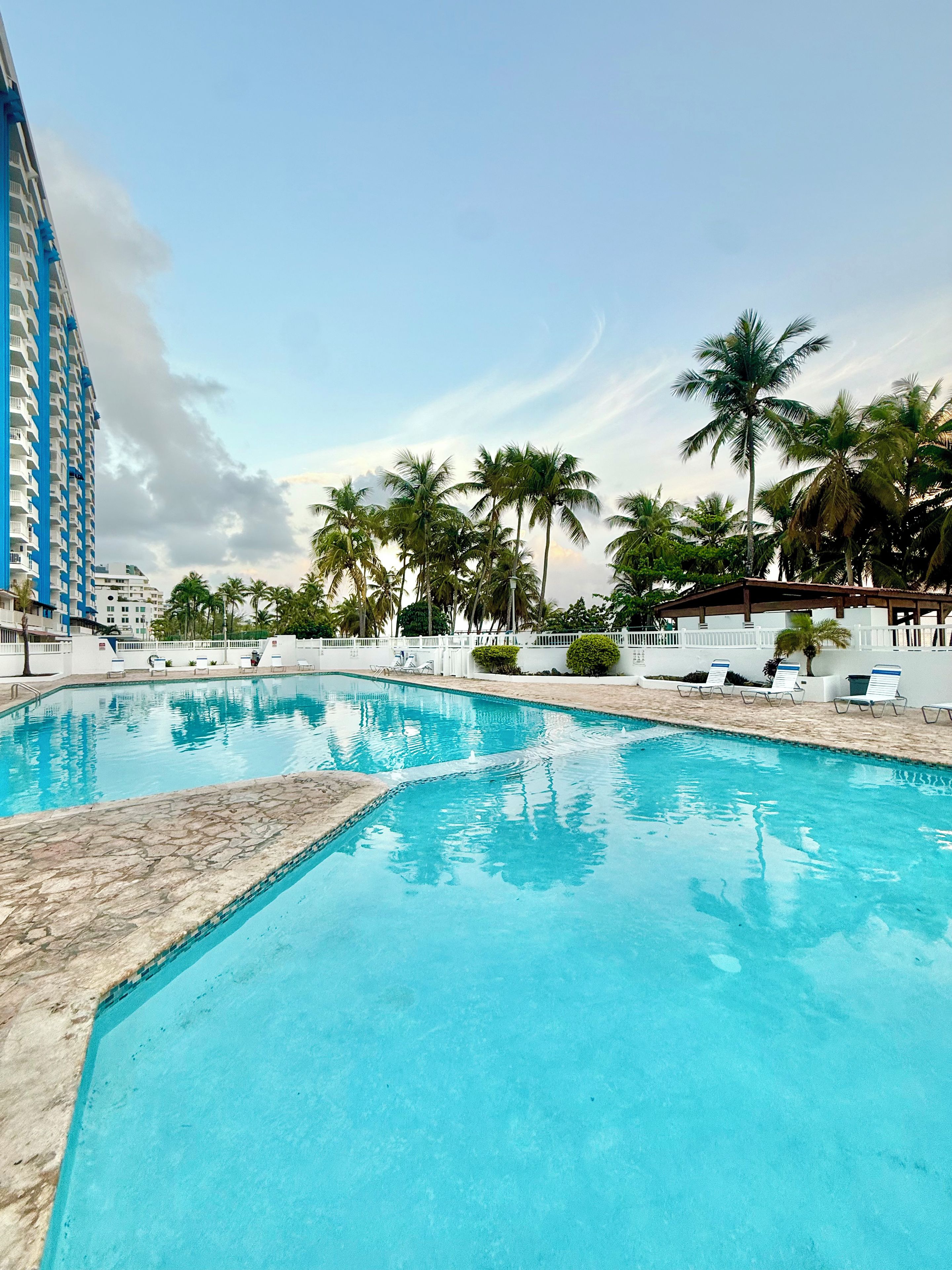 A swimming pool with clear blue water, surrounded by palm trees and a white building with blue accents.