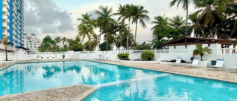 Ein Pool mit klarem, blauem Wasser, umgeben von Palmen und einem weißen Gebäude mit blauen Akzenten.