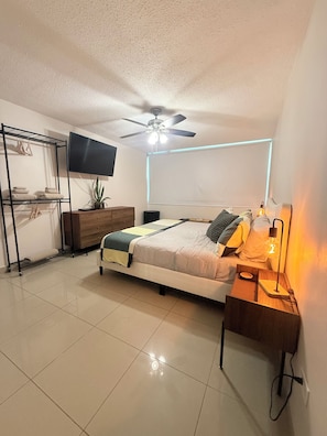 A bedroom with a bed, a ceiling fan, a TV, a bedside lamp, and a wall-mounted shelf. - Brisa Caribe Marbella Elegant 3BR Retreat Steps from Beach in Isla Verde, PR!! (Carolina)