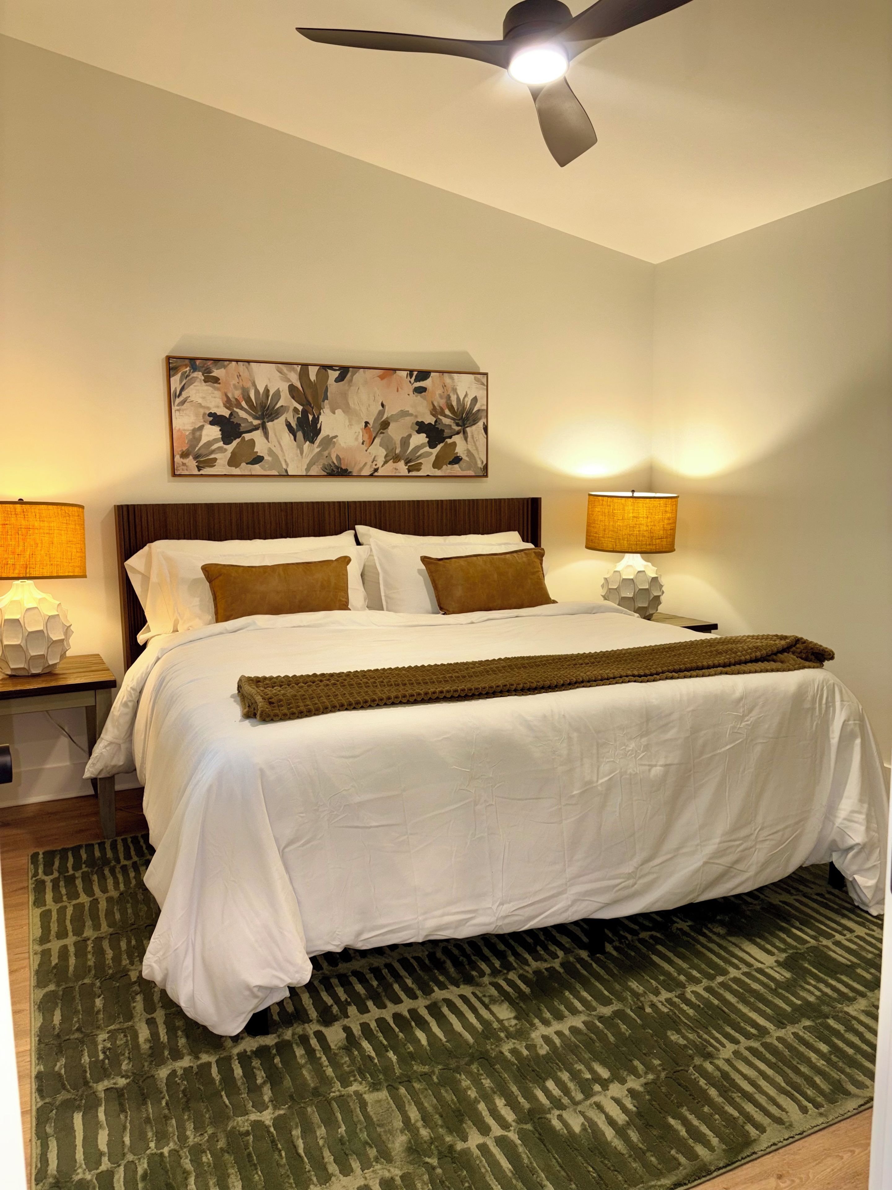 Habitación de hotel con cama, dos lámparas de noche, ventilador de techo, obra de arte en la pared y alfombra estampada.
