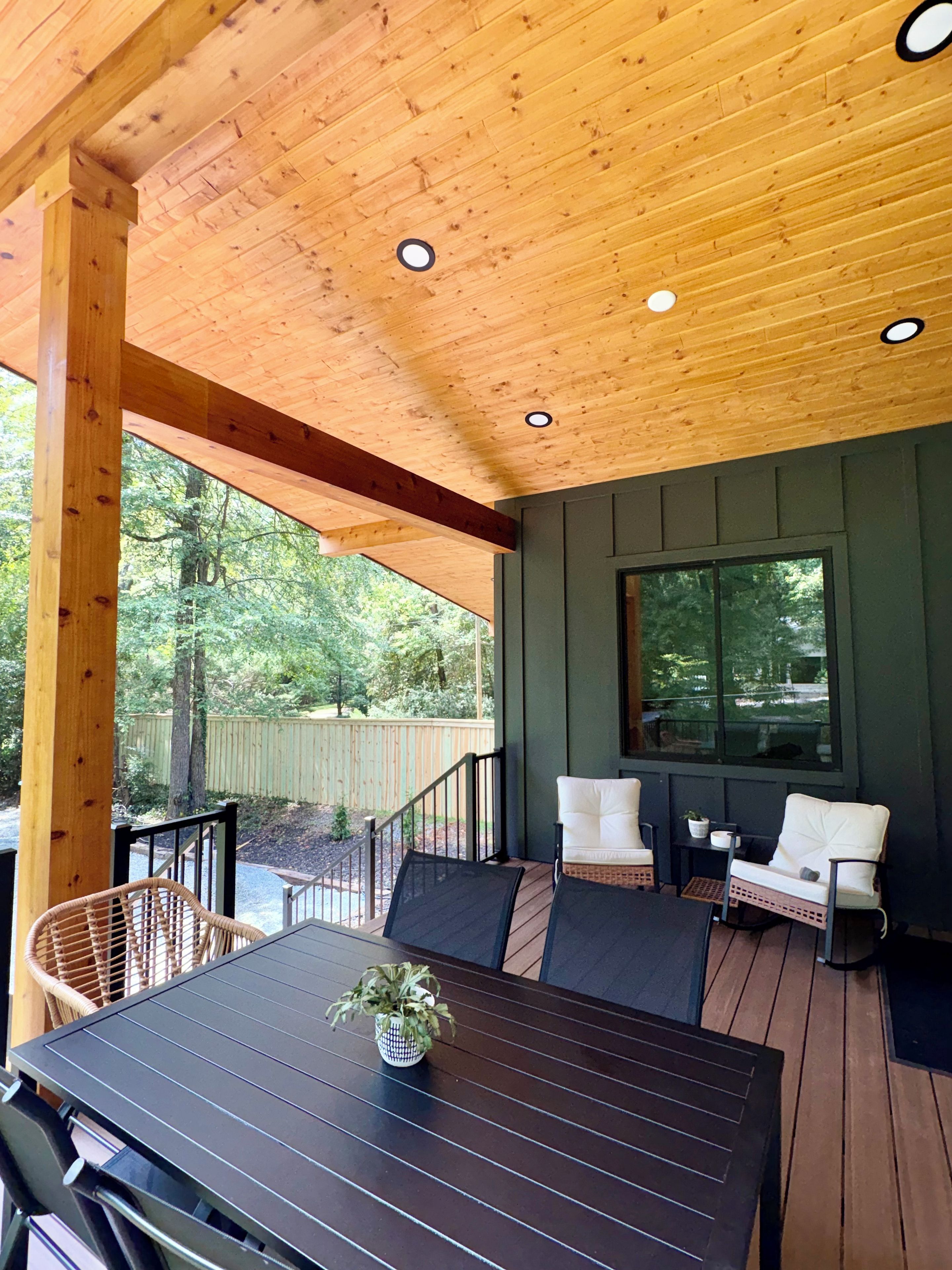 A covered outdoor seating area with a table and chairs, a window, and a view of the surrounding trees.