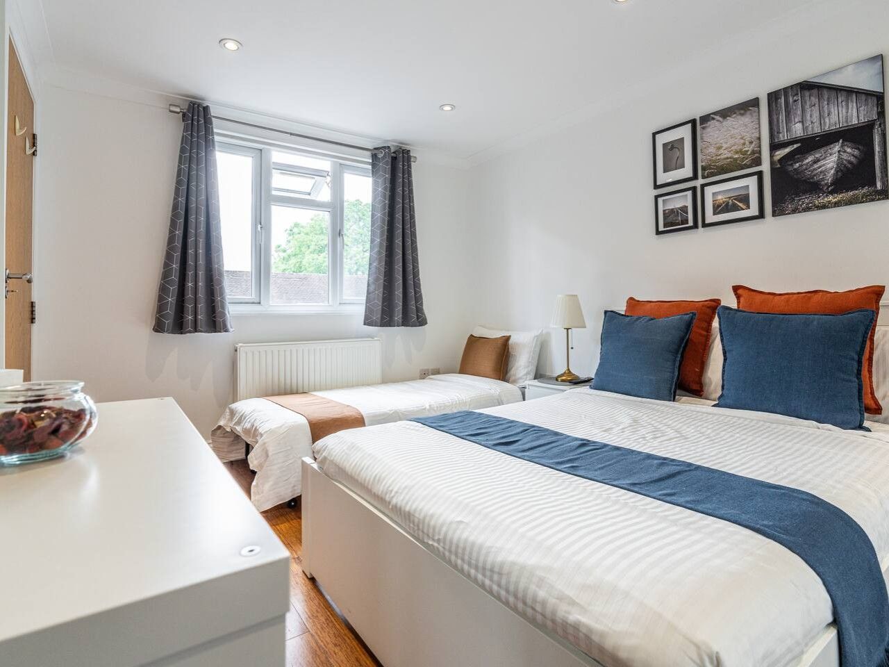 A hotel room with two beds, a white desk, a bowl of fruit, and a window with curtains.